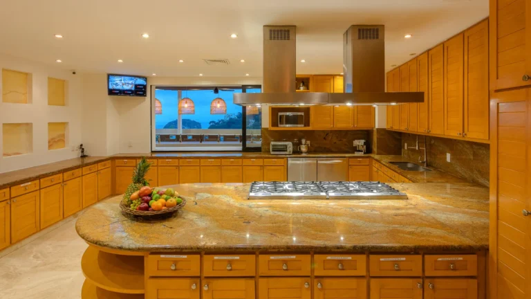 Villa Magnifico Kitchen with wooden cabinets, granite island, fruit basket; ocean-view windows. Mismaloya luxury villa near Puerto Vallarta, Mexico.