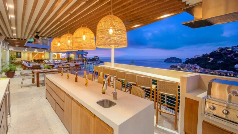 Villa Magnifico Outdoor kitchen/bar in Mismaloya, near Puerto Vallarta: modern wood cabinetry, wicker-lit pendants, stools, overlooking bay at dusk.