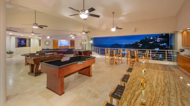 Villa Magnifico Indoor game room with tables, bar seating; high ceilings, windowed wall displaying Mismaloya's coastal cityscape and hills.