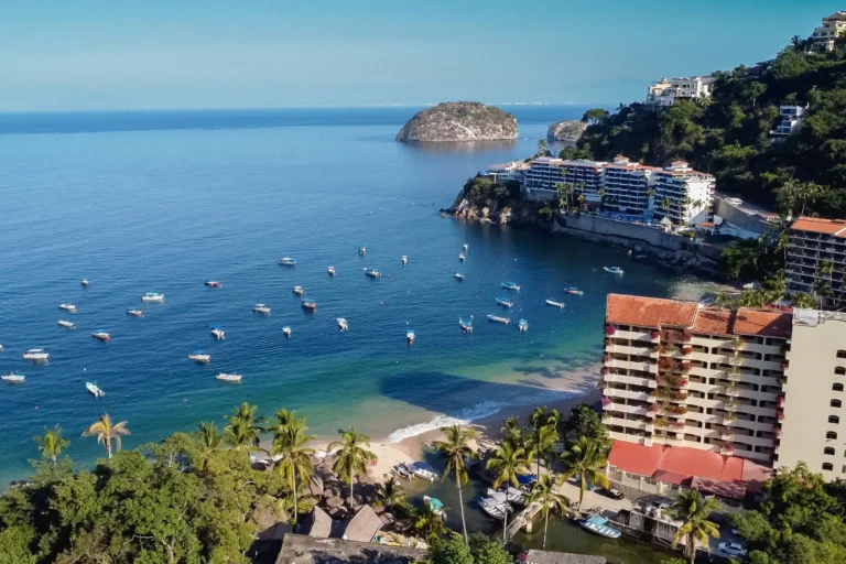 Villa Magnifico Coastal view of Mismaloya, near Puerto Vallarta: palm-lined beach, anchored boats, hillside red-roof villas overlooking the ocean.