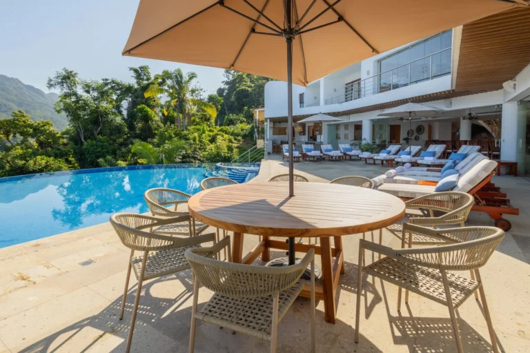 Villa Magnifico Outdoor patio in Mismaloya: wooden table, wicker chairs under umbrella. Infinity pool, lounge chairs, overlooking lush hills.